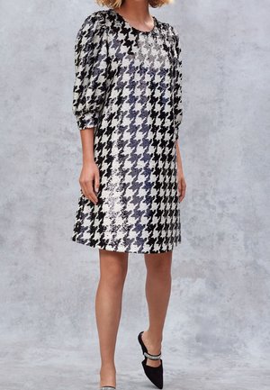 Woman wearing knee-length black and white houndstooth sequin dress with puffed three-quarter sleeves and black pointed-toe heels with rhinestone straps.