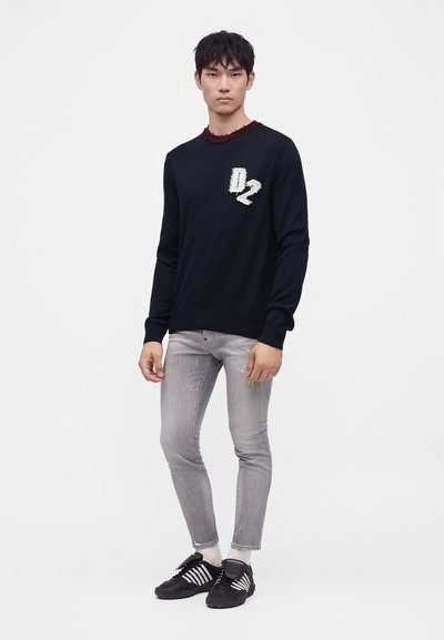 Jeune homme portant un pull noir avec un patch "D2", un jean skinny gris clair déchiré et des baskets noires à rayures blanches, debout sur un fond blanc.