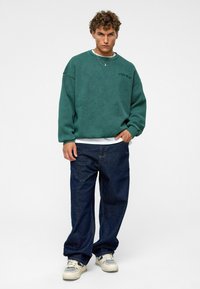 Teal oversized sweatshirt met verlaagde schouders, gecombineerd met losse donkerblauwe jeans. Witte onderhemd zichtbaar aan de onderkant, en beige sneakers.