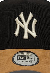 Gorra negra con un logotipo bordado en blanco de "NY". Presenta una visera marrón y una correa ajustable. Incluye la marca New Era en la parte interior.