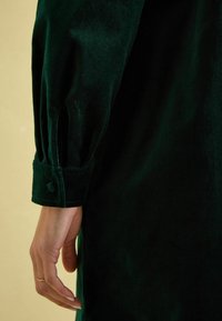 Grünes Samtshirt mit langen Ärmeln, ausgestattet mit geknöpften Manschetten und einer weichen Textur, die durch einen subtilen Schimmer vor einem schlichten Hintergrund hervorgehoben wird.