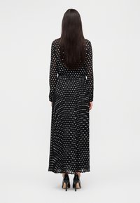 Μάξι φόρεμα με μαύρα πουά, μακριά μανίκια, στενή μέση και ρέοντα φούστα. Φορεμένο με μαύρα παπούτσια με τακούνι, εμφανίζεται από πίσω.