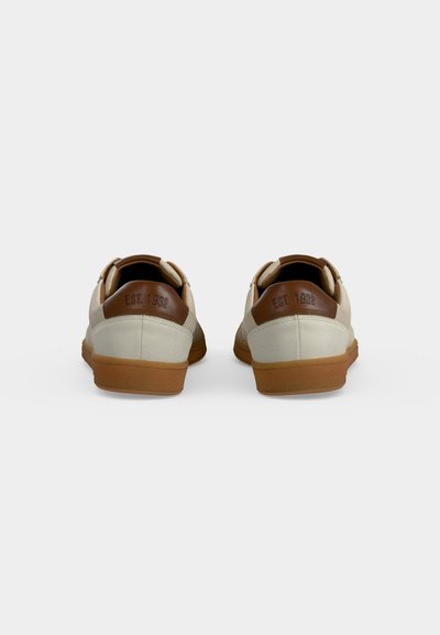 Paire de baskets beige et marron avec semelles en gomme vue de dos, avec « EST. 1932 » embossé sur les languettes marron du talon.
