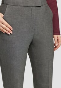 Graue, maßgeschneiderte Hose aus strukturiertem Stoff, mit Fronttaschen und einem glatten Bund, gezeigt mit einer Hand, die auf dem Oberschenkel ruht.