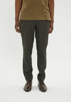Mężczyzna w ciemnoszarych, szytych na miarę spodniach, brązowych skórzanych sznurowanych butach i oliwkowozielonym t-shircie, stojący na jednolitym tle.
