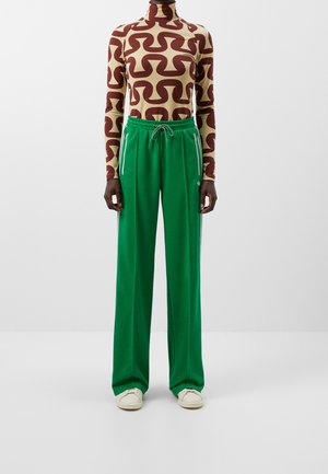 Persoon draagt een beige en bruin patroon coltrui, felgroene losse broek met trekkoord en ritsen, en witte sneakers, tegen een effen achtergrond.