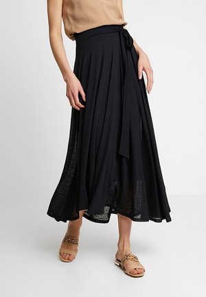 Femme portant une longue jupe noire fluide nouée à la taille, un top beige sans manches et des sandales plates beiges avec des brides à motifs.