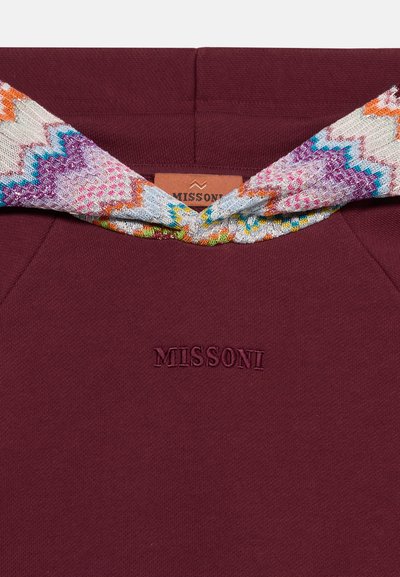 Μποργκουντί ύφασμα με κεντημένο λογότυπο Missoni και Πολύχρωμο πλεκτό φουλάρι με ζιγκ-ζαγκ σχέδιο δεμένο στην κορυφή.