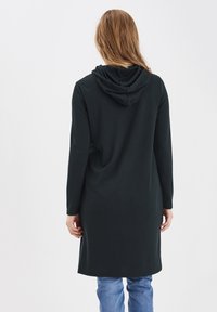 Lång svart hoodieklänning av mjukt material, med långa ärmar, en huva bak och rak nederkant. Bärs med ljusblå jeans.