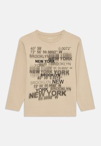 Beige Langarm-T-Shirt mit schwarzem Aufdruck von Städtenamen und geografischen Koordinaten in verschiedenen Schriftarten und -größen.