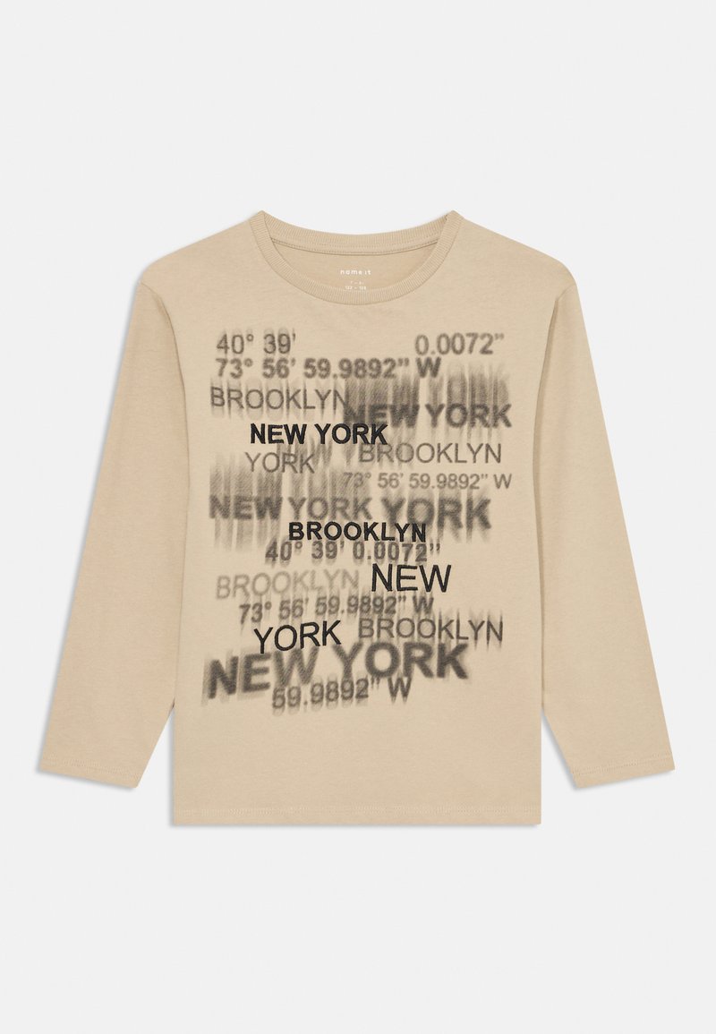 Hosszú ujjú bézs póló, fekete nyomtatott szöveggel, amely városneveket és földrajzi koordinátákat tartalmaz különböző betűtípusokban és méretekben.