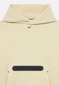 Hanorac bej cu glugă mare, logo Nike negru și o buzunar frontal proeminent cu fermoar și margine neagră. Textură de material moale și neted.