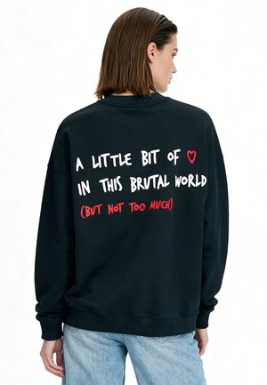 Personne portant un sweat-shirt noir avec un texte au dos indiquant "A little bit of heart in this brutal world (but not too much)".