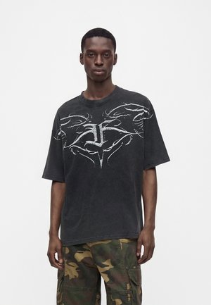 Mand iført en sort oversized t-shirt med hvidt abstrakt grafik og camouflage cargobukser, der står mod en ensfarvet hvid baggrund.