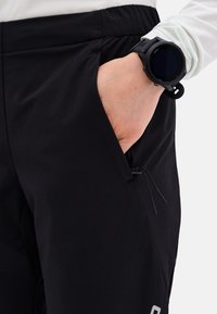 Schwarze Hose aus strukturiertem Stoff, mit Seitentaschen und Kordelzugdetails. Am Handgelenk befindet sich eine schwarze Uhr mit rundem Ziffernblatt und digitaler Anzeige.