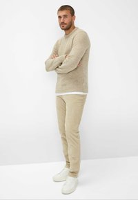 Beige gebreide trui met geribde manchetten en zoom, gecombineerd met lichtbeige corduroypants en witte sneakers, staande tegen een neutrale achtergrond.