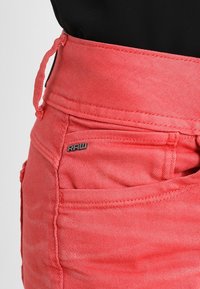 Falda de mezclilla coral con cintura alta, que presenta un cordón y bolsillos laterales. Destaca el acento del logo "RAW" en la parte frontal. La textura es suave.
