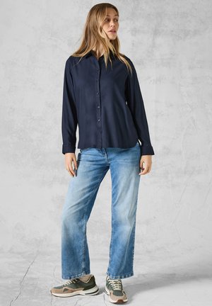 Donkerblauwe button-upshirt met lange mouwen, gecombineerd met lichtblauwe wijde jeans en grijs-groene sneakers met crèmekleurige accenten.