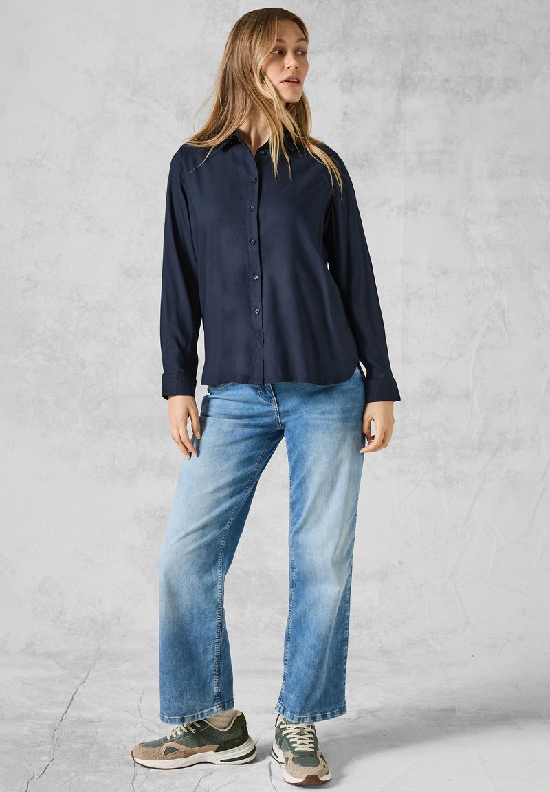 Camicetta blu scuro con bottoni e maniche lunghe, abbinata a jeans a gamba larga blu chiaro e scarpe da ginnastica grigio-verdi con accenti crema.