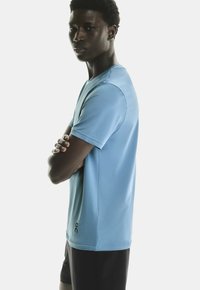 T-shirt de sport bleu clair à manches courtes avec un col rond, texture lisse et un logo imprimé près de l'ourlet. Porté avec un short sombre.