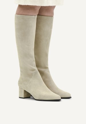 Knielange beige Wildlederstiefel mit Blockabsätzen, getragen zu einem weißen Rock vor hellem Hintergrund.