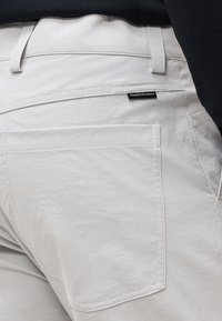 Gros plan d'une poche de pantalon beige clair avec des coutures visibles et une petite étiquette noire « Peak Performance » sur la poche arrière.