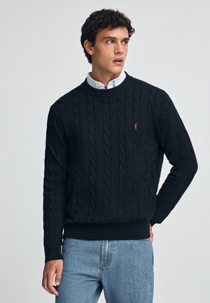 Zwarte kabelgebreide trui met een ronde halslijn en een rood logoaccent. Draag het over een lichtblauw overhemd met kraag, gecombineerd met lichte jeans.