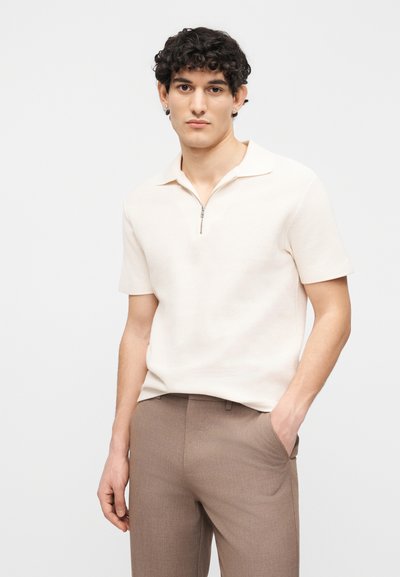 Άνδρας με σγουρά μαλλιά, ντυμένος με κοντομάνικο πλεκτό πόλο σε κρεμ απόχρωση με φερμουάρ και ανοιχτό καφέ παντελόνι, που στέκεται με το ένα χέρι στην τσέπη.