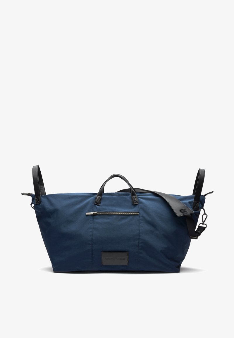 Borsa da viaggio blu navy realizzata in tessuto resistente con dettagli in pelle nera, tasca con zip, doppi manici e tracolla regolabile.