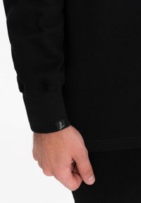 Musta pitkähihainen paita, jossa on teksturoitu kangas, istuvat hihansuut ja pieni valkoinen etiketti lähellä rannetta. Läheltä otettu kuva kädestä, joka pitää hihasta kiinni.