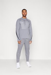 Graues Sportoutfit mit einem Langarmoberteil mit gestepptem Frontpanel und einer schlanken Passform für die Unterteile. Akzente in Schwarz. Die Schuhe sind weiß.
