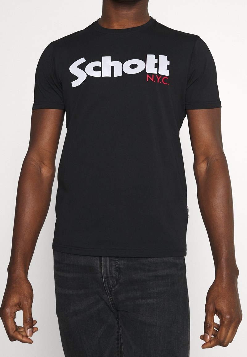 Personne portant un t-shirt noir à manches courtes avec un texte blanc "Schott" et un texte rouge plus petit "N.Y.C." sur la poitrine, associé à un jean foncé.