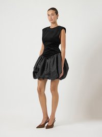 Vestido preto com corpete justo e saia volumosa e franzida. Feito de um tecido texturizado, combinado com sapatos de salto fino, de ponta arredondada, em tom castanho.