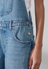 Prim-plan cu o persoană ce poartă salopetă denim albastru deschis cu nasturi metalici și o cămașă albă, arătând buzunarul lateral și brațul.