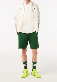 Person som har på sig en vit zip-jacka, vit polo-tröja, mörkgröna shorts, gröna strumpor med vita ränder och neon gula sneakers, står med händerna i fickorna.