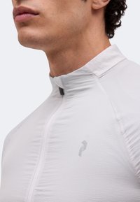 Κοντινό πλάνο στο κάτω μέρος του προσώπου και στον λαιμό ενός άνδρα που φοράει ανοιχτογλογέ γκρι αθλητικό μπλουζάκι με φερμουάρ και μικρό λογότυπο στο στήθος.