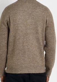 Personne portant un pull marron tricoté à manches longues avec un ourlet côtelé, vue de dos sur un fond uni.