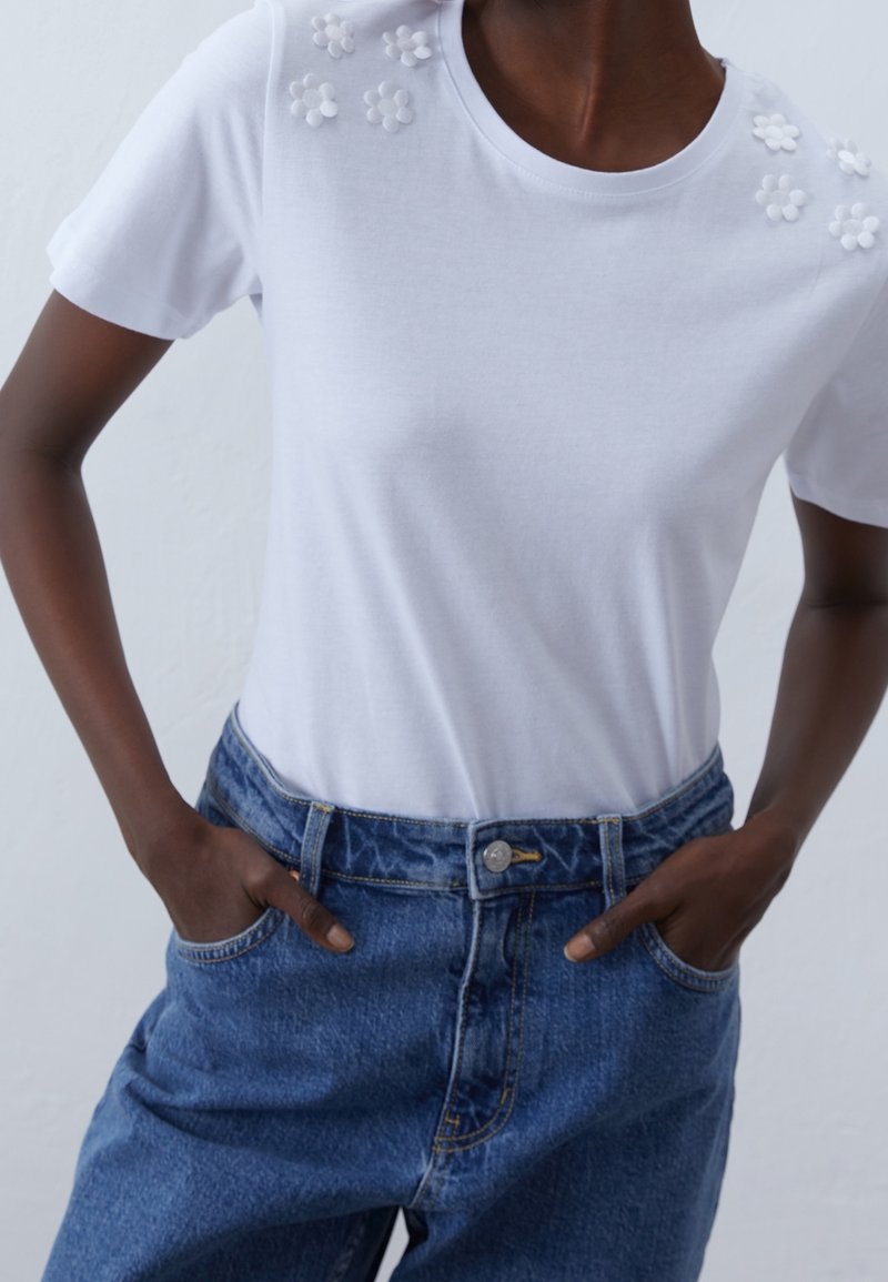 Personne portant un haut blanc à manches courtes avec de petits motifs floraux en 3D sur les épaules et un jean bleu taille haute avec les mains dans les poches.