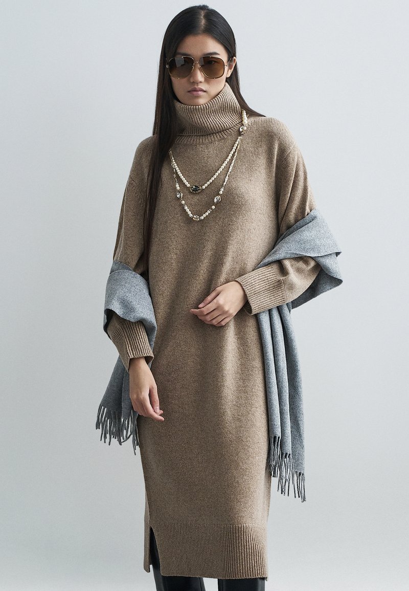 Smeđa pletena haljina s rolkom, s rebrastim rubom i bočnim prorezima, slojevito nošena sa sivim šalom, ukrašena višebojnom ogrlicom i sunčanim naočalama.