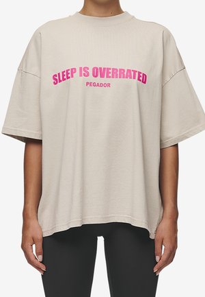 Beżowy oversize t-shirt z różowym napisem „SLEEP IS OVERRATED” i mniejszym „PEGADOR” noszony przez osobę w czarnych spodniach.