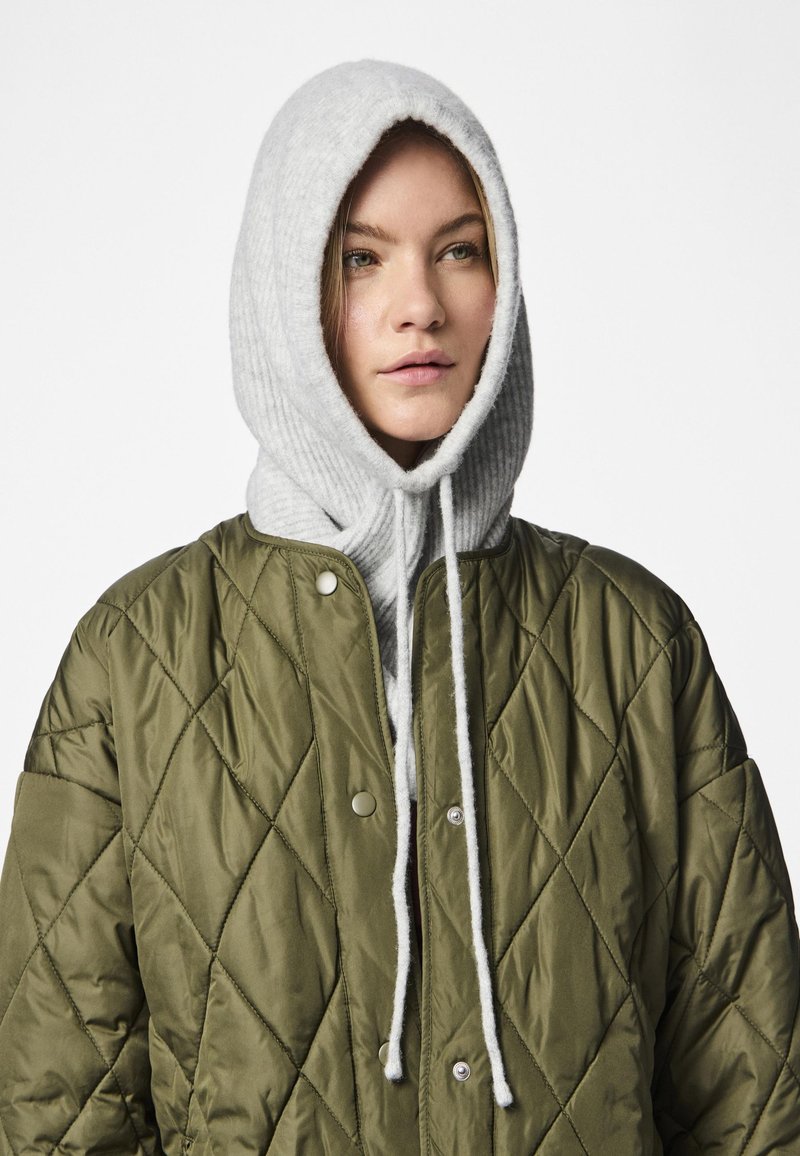 Giacca trapuntata verde oliva con bottoni a pressione argentati, sovrapposta a un cappuccio grigio chiaro a coste, caratterizzata da lacci regolabili e una texture morbida.