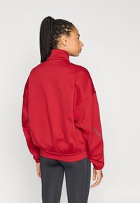 Červená mikina s vysokým golierom, nadmerným strihom, rebrovanými manžetami a pásom. Má hladkú textúru a kontrastné sivé detaily.