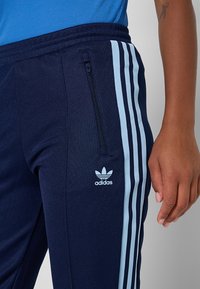 Námornícky modré teplákové nohavice Adidas s svetlomodrými pruhmi. Majú vrecko so zipsom, rebrovaný pás a malú značku na ľavom stehne.