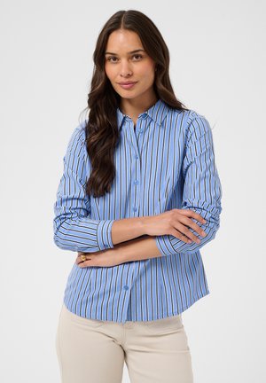Femme aux longs cheveux foncés portant une chemise à rayures verticales bleues et blanches avec boutons et un pantalon beige clair, bras croisés, fond neutre.