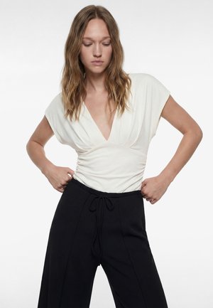 Frau mit welligem Haar, die ein weißes Top mit tiefem V-Ausschnitt und hoch taillierte schwarze Hose trägt und die Hose an der Taille auf einem weißen Hintergrund richtet.