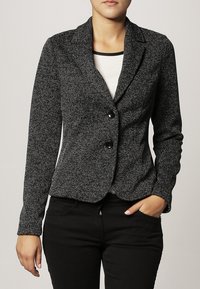 Blazer texturé noir et blanc avec col cranté, deux boutons, manches longues et coupe ajustée, associé à un haut blanc et un pantalon noir.