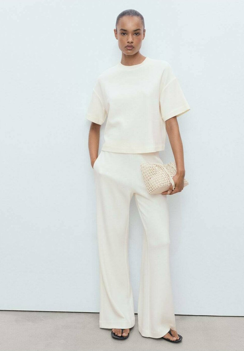 Set coordinato color panna composto da una blusa corta a maniche corte e pantaloni a gamba larga, abbinato a una clutch a trama color beige chiaro e sandali neri.