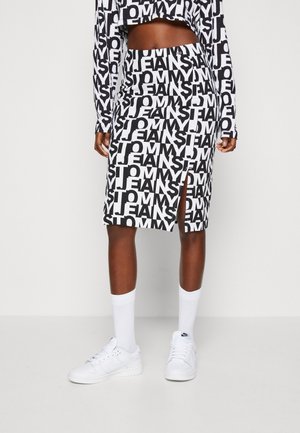 Vrouw draagt een zwart-witte rok met patroon en een bijpassende top met lange mouwen, witte sneakers en sokken, staand tegen een effen achtergrond.