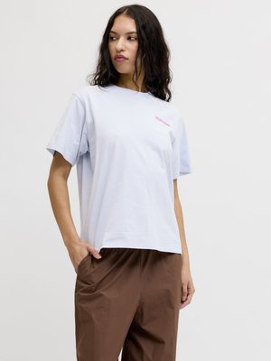 Femme aux cheveux bruns ondulés portant un t-shirt bleu clair avec le texte "WILD FLOWER" et un pantalon marron, debout avec une main dans la poche.