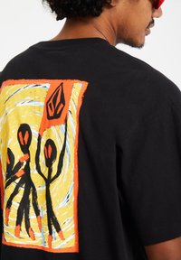 Black cotton t-shirt featuring a colorful graphic design with abstract figures in orange, black, and yellow on the back.